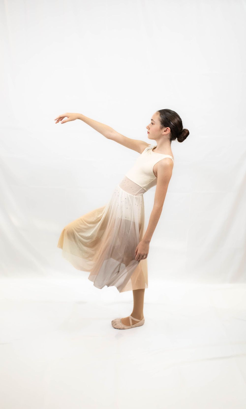 Ballet dancer in flowing beige dress strikes graceful pose against white background.