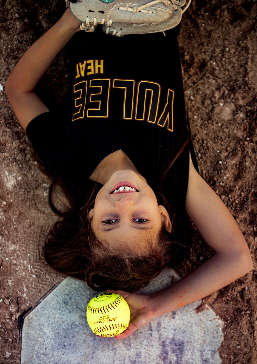 Softball player in black jersey lying on ground holding bright yellow softball in creative portrait series.