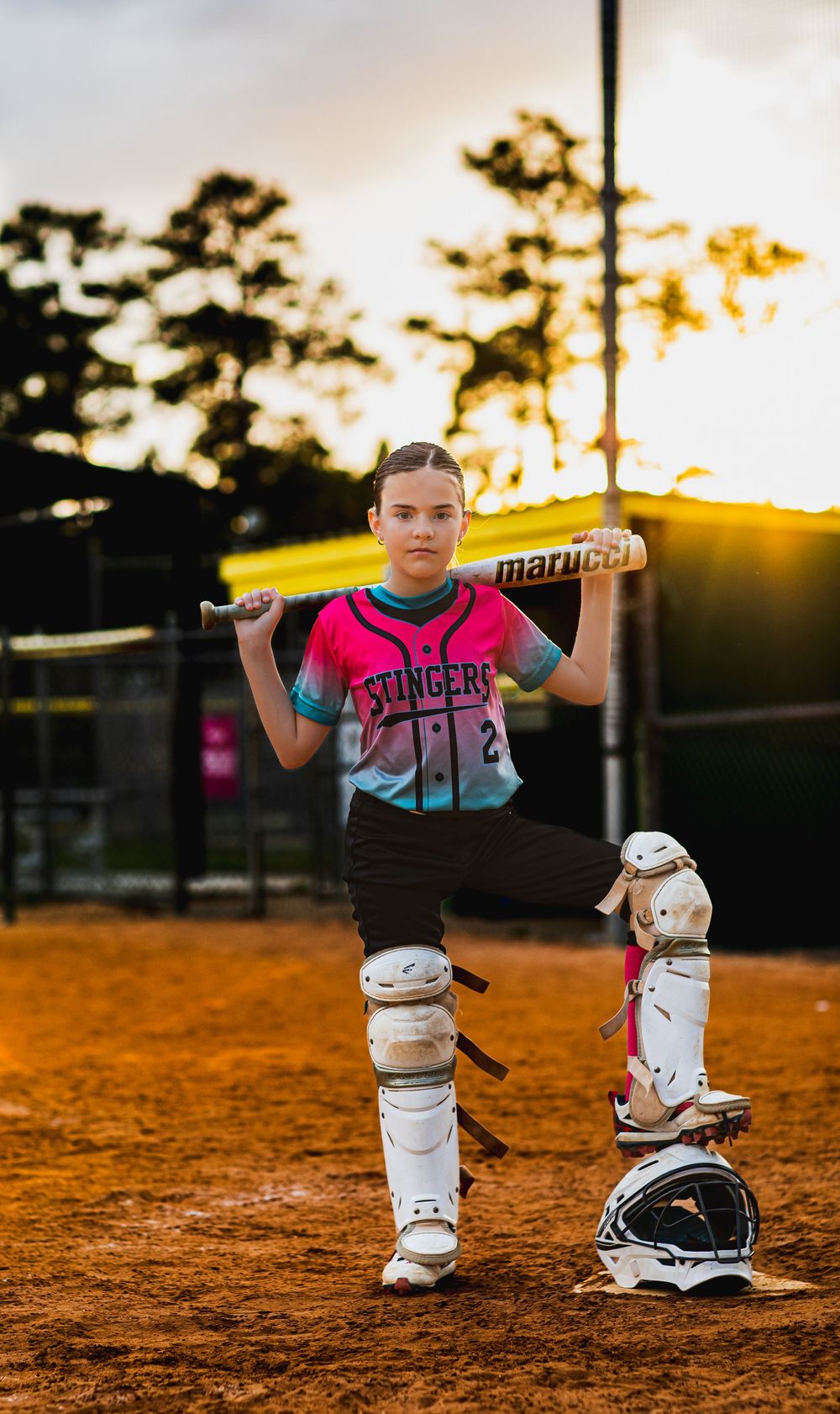 Young softball player in pink Stingers jersey holds Marucci bat on shoulders wearing catcher gear at sunset field.