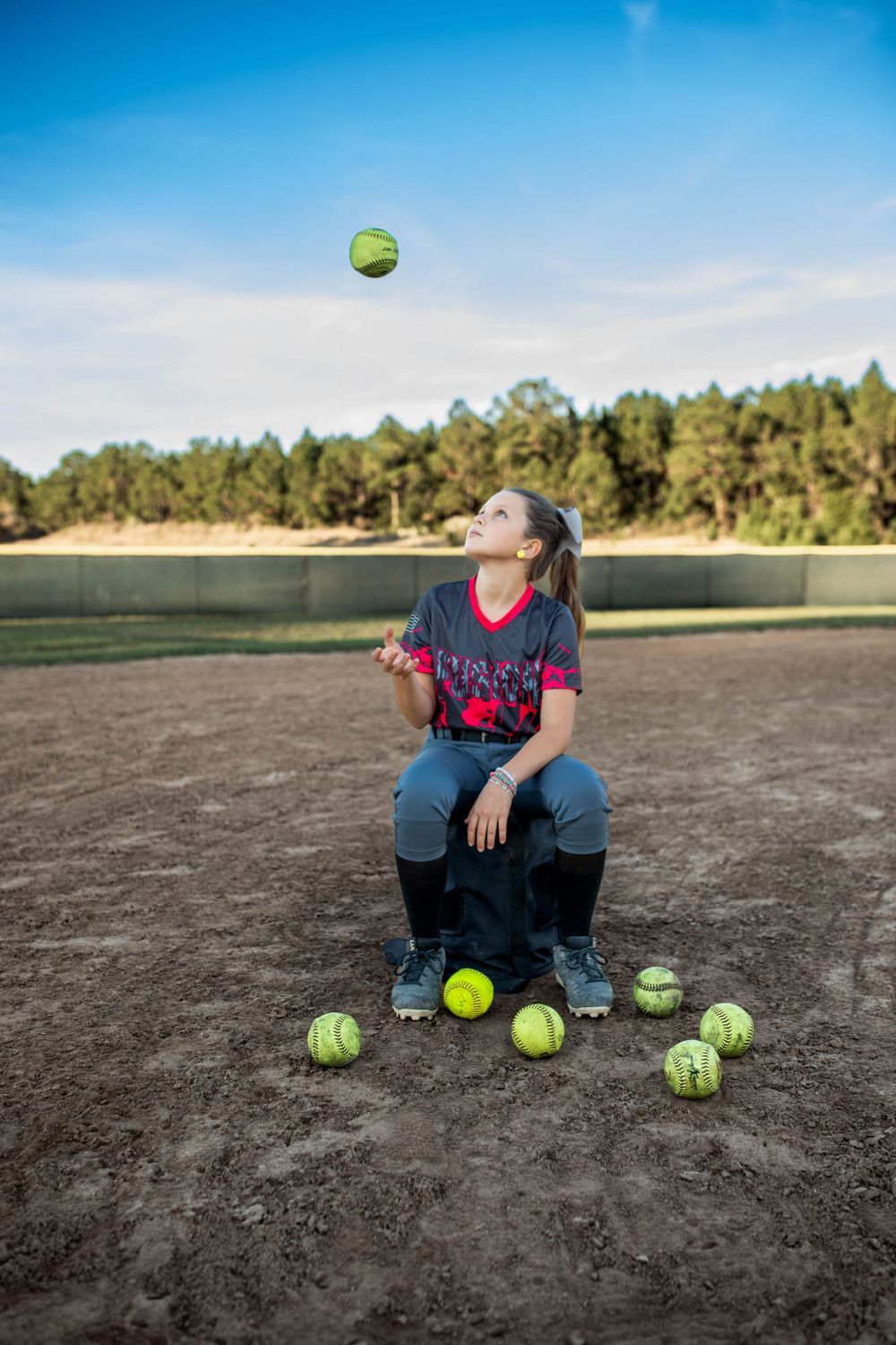 Softball player sitting on field with multiple softballs scattered around while one ball floats above on sunny day.
