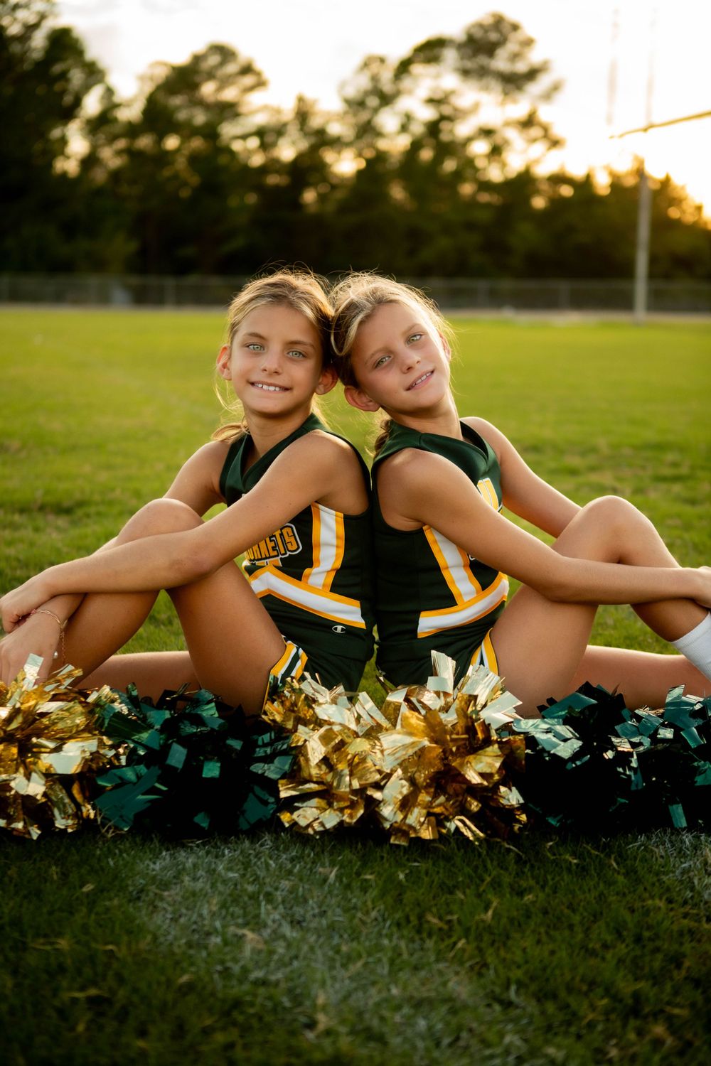 Two cheerleaders in matching uniforms pose back-to-back on sports field at sunset.
