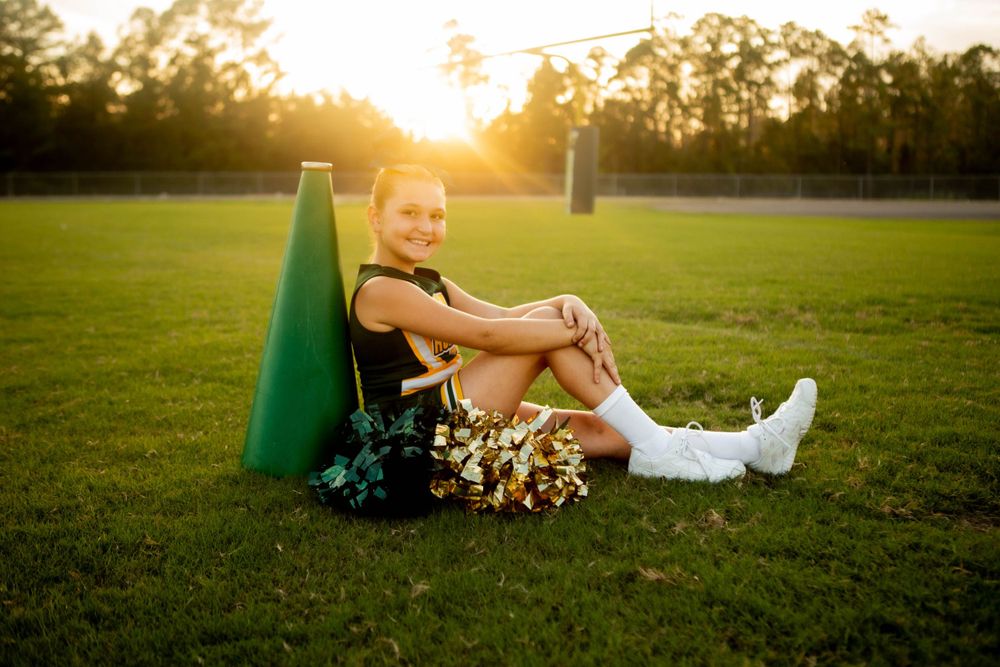 A cheerleader in uniform sits on a football field at sunset with pom poms and athletic gear.