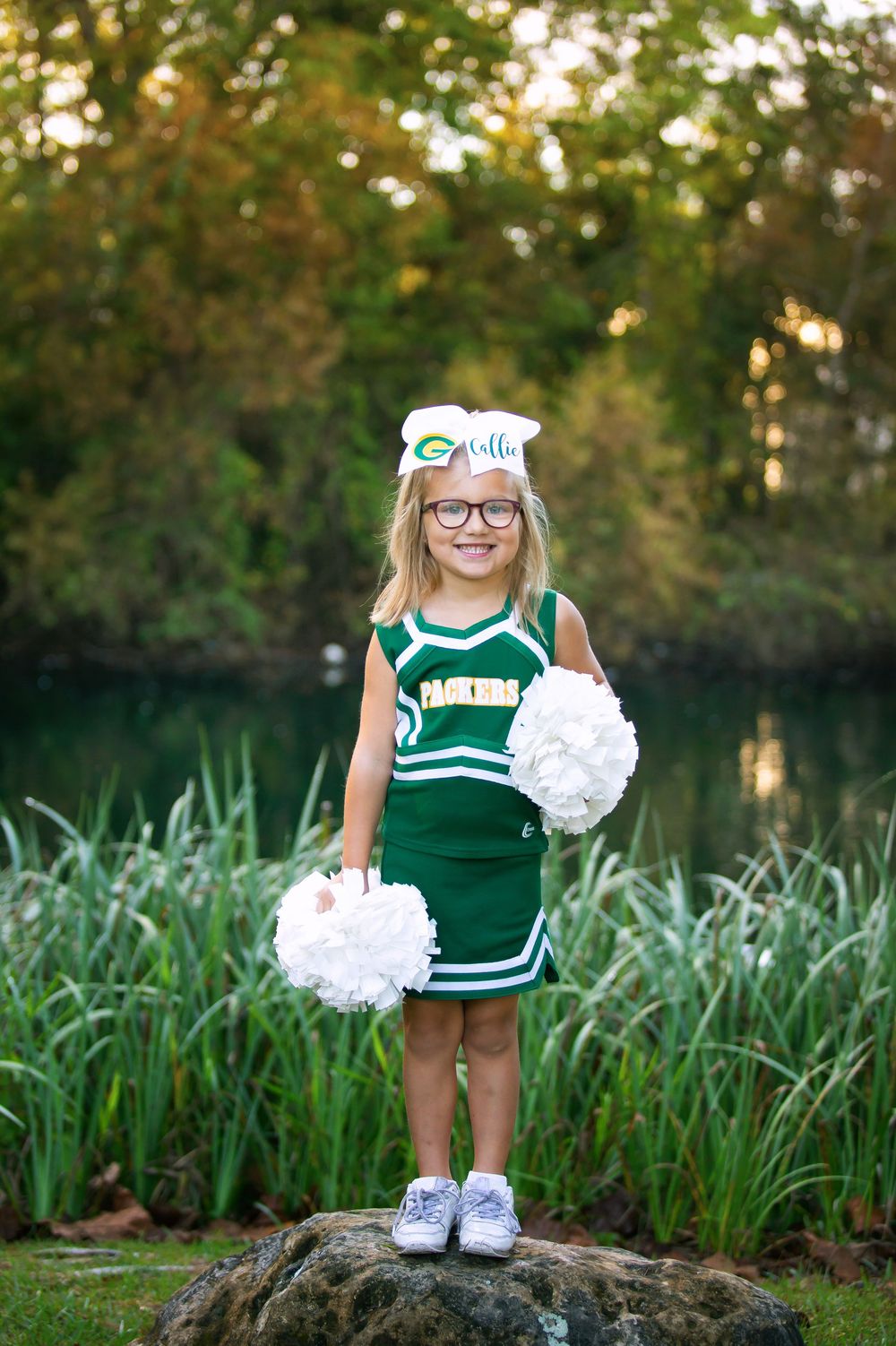 A cheerleader in green and white uniform poses with pom poms in a garden setting during golden hour.