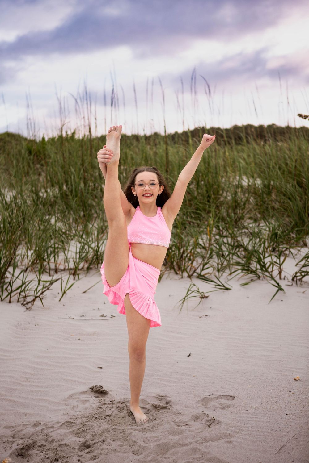Dancer in pink outfit performs graceful pose on sandy beach with beach grass and cloudy sky in background.