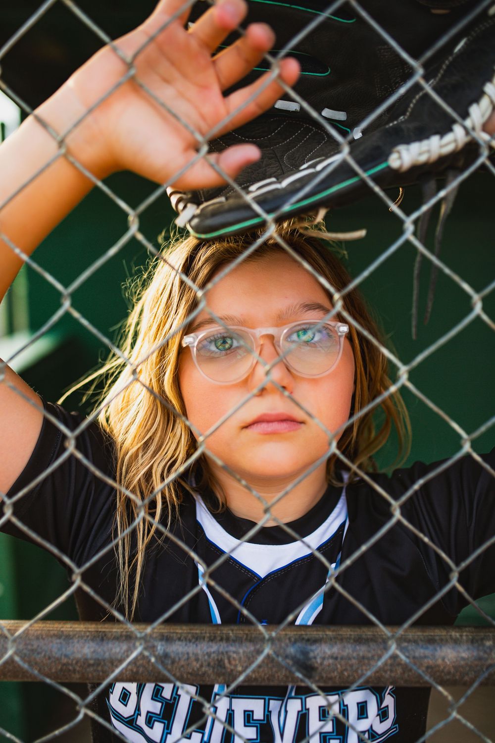 Young softball player in Believers jersey and glasses leaning against chain-link fence dugout, looking intensely at camera.
