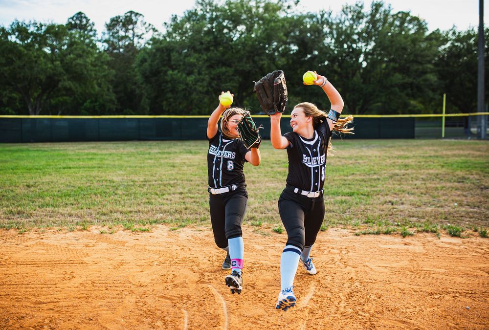 Two softball players in black uniforms celebrate on the field, each holding a softball up in the air.