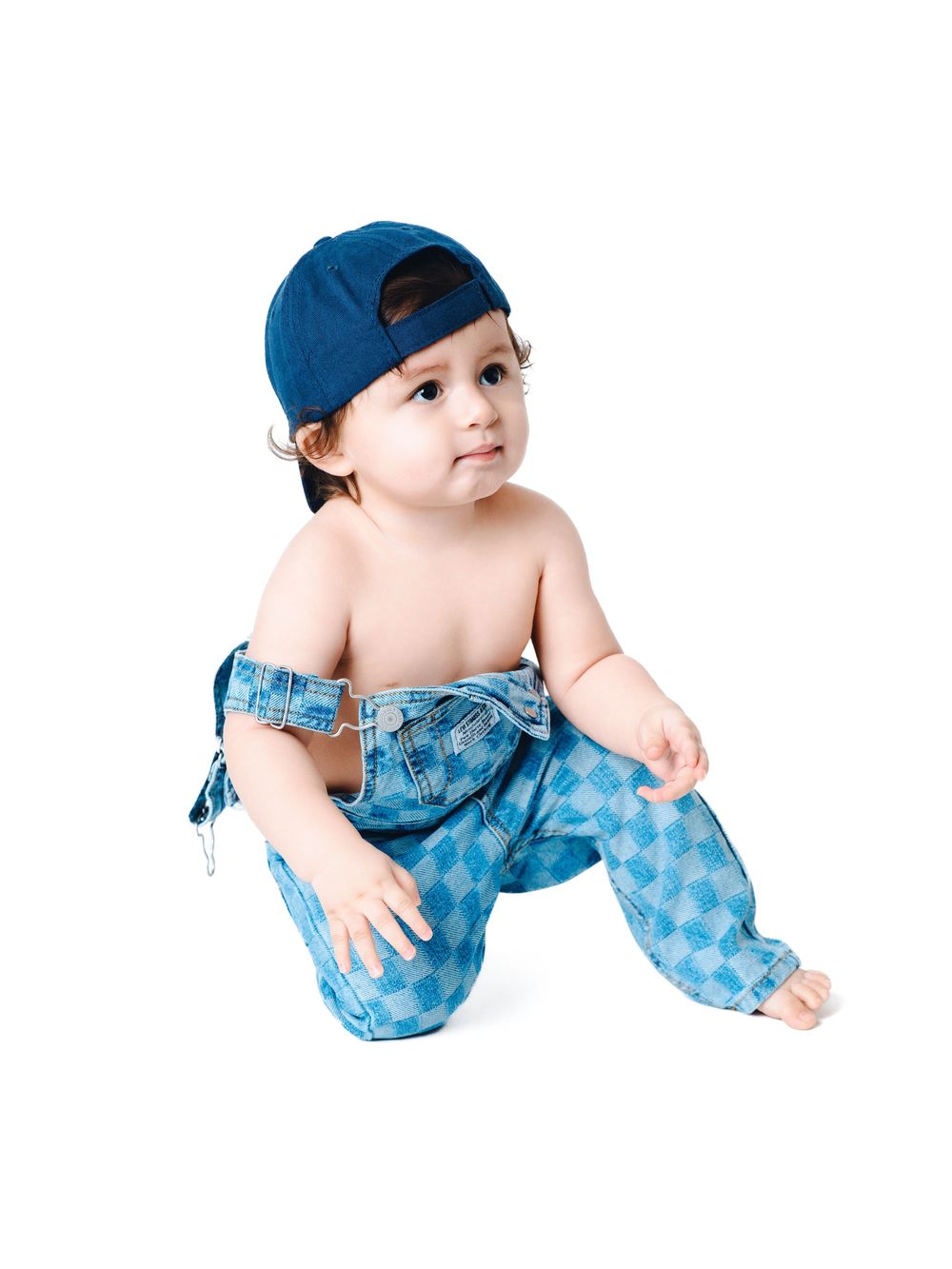 Cute baby in blue checkered overalls and backwards cap kneeling on white background.