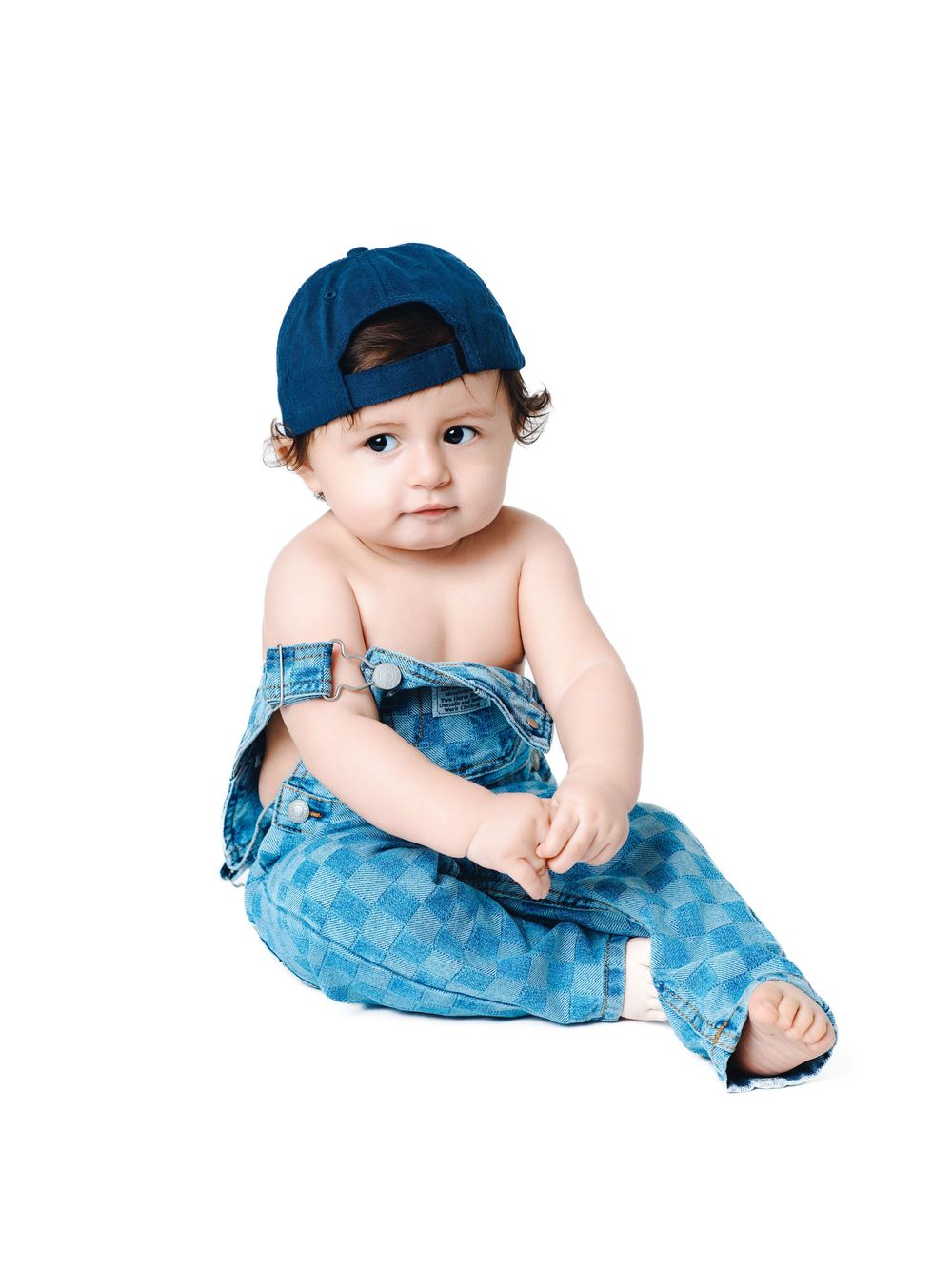 Cute baby wearing blue denim overalls and a backwards baseball cap sitting on a white background.
