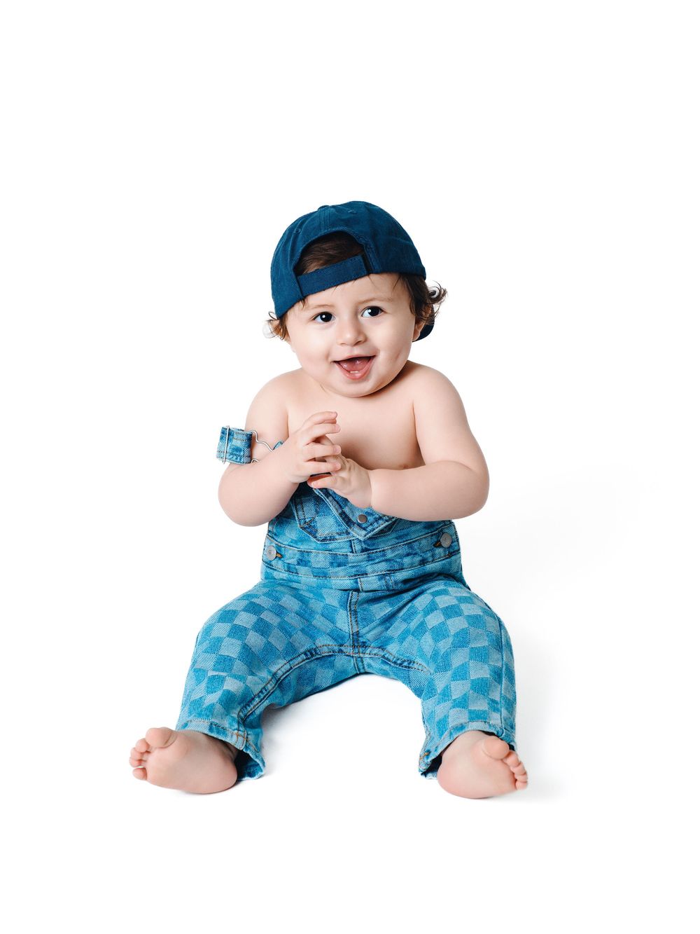 Smiling baby in blue checkered overalls and backwards cap sitting on white background.