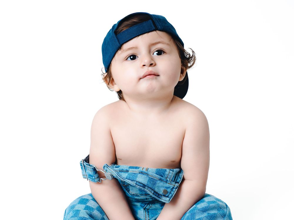 Cute toddler boy wearing a blue baseball cap and denim shorts, sitting against a white background.