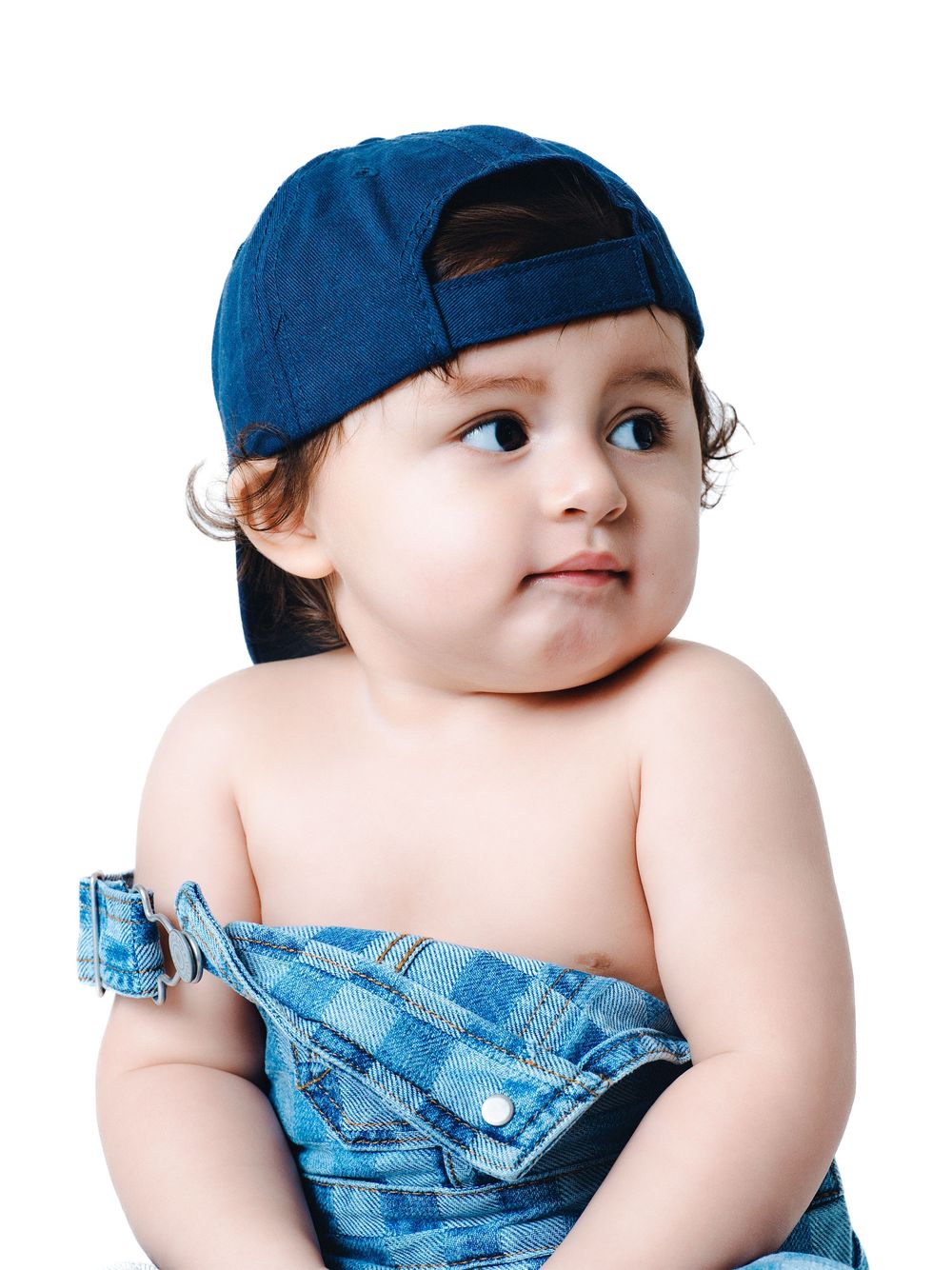 Cute toddler wearing a blue denim cap backwards and denim overalls, looking sideways on white background.