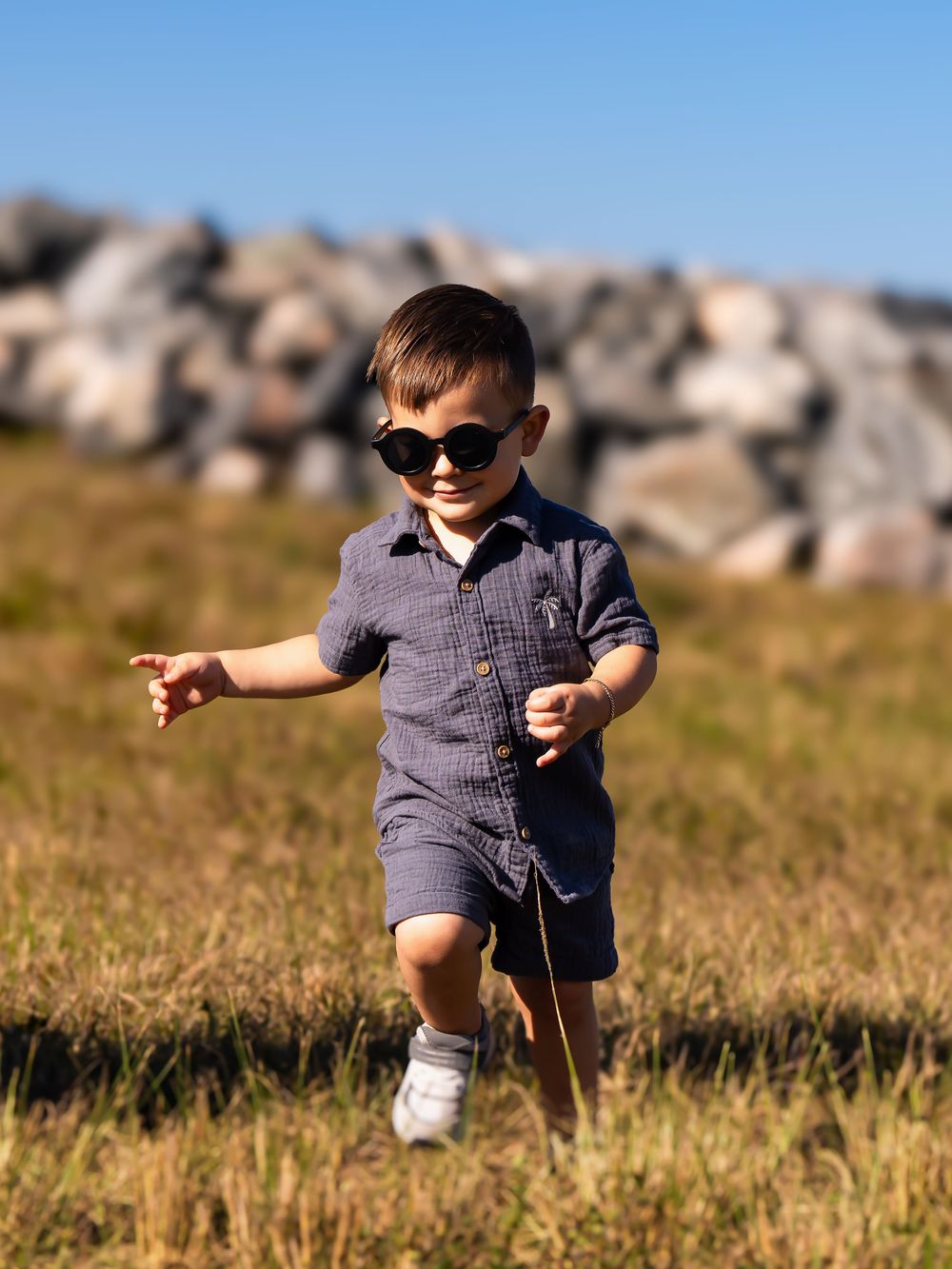 A cheerful toddler boy in navy shorts set and sunglasses runs through a grassy field with rocky hills in the background.