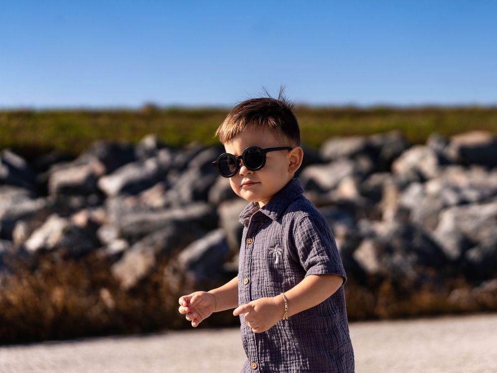 A stylish toddler boy wearing black sunglasses and a blue plaid shirt stands outdoors near rocky terrain.
