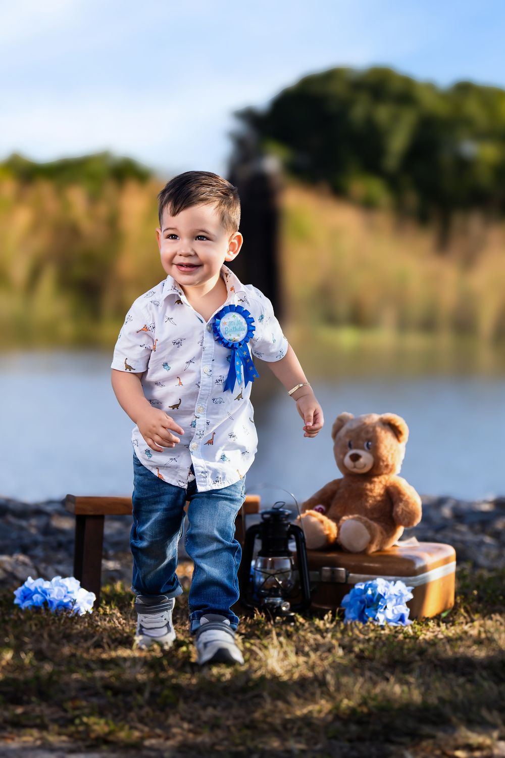 Smiling toddler boy in white shirt and jeans with blue ribbon badge stands outdoors near teddy bear and vintage suitcase.