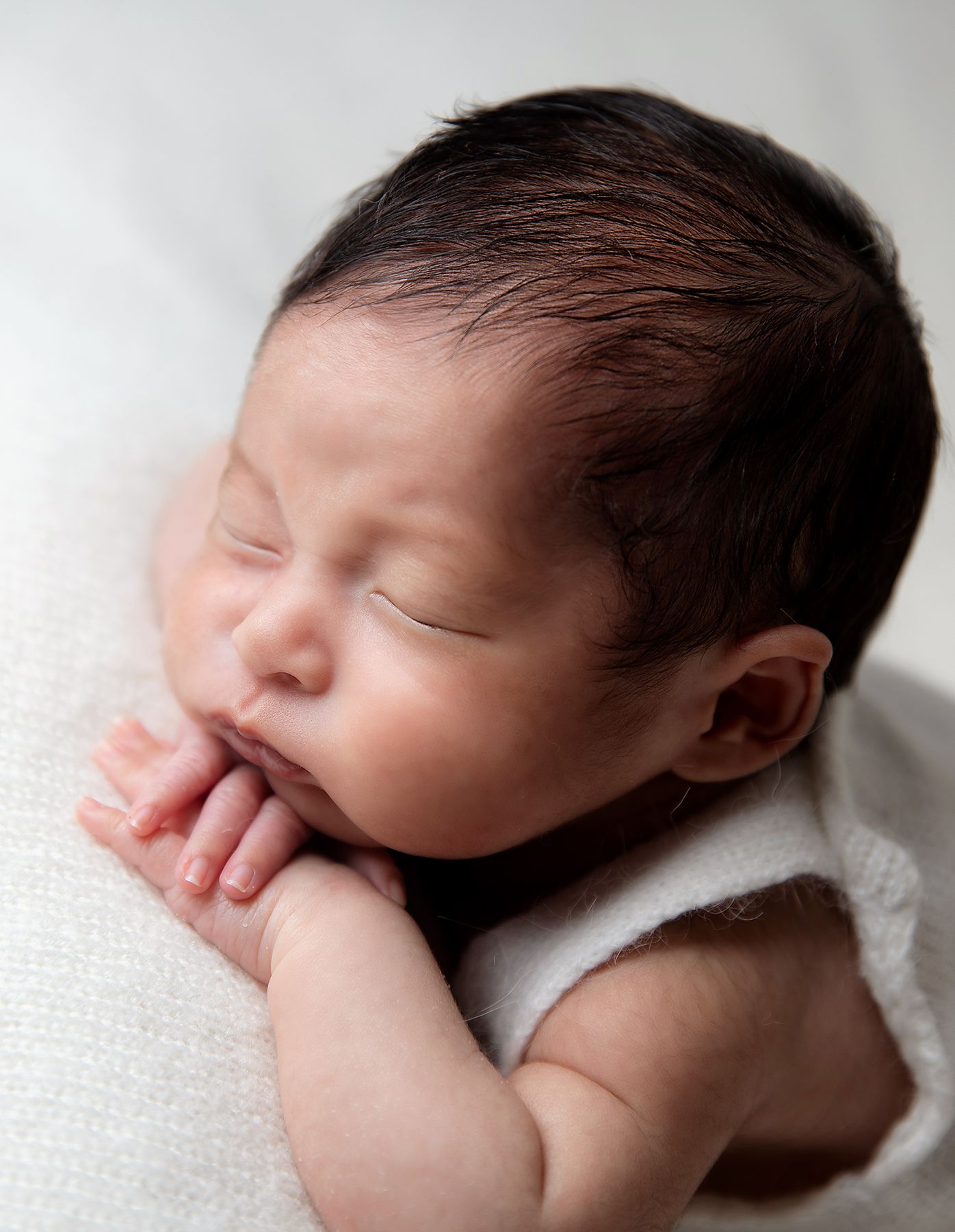 Peaceful sleeping newborn baby wrapped in white blanket with hand near face against light background. During a photoshoot in London