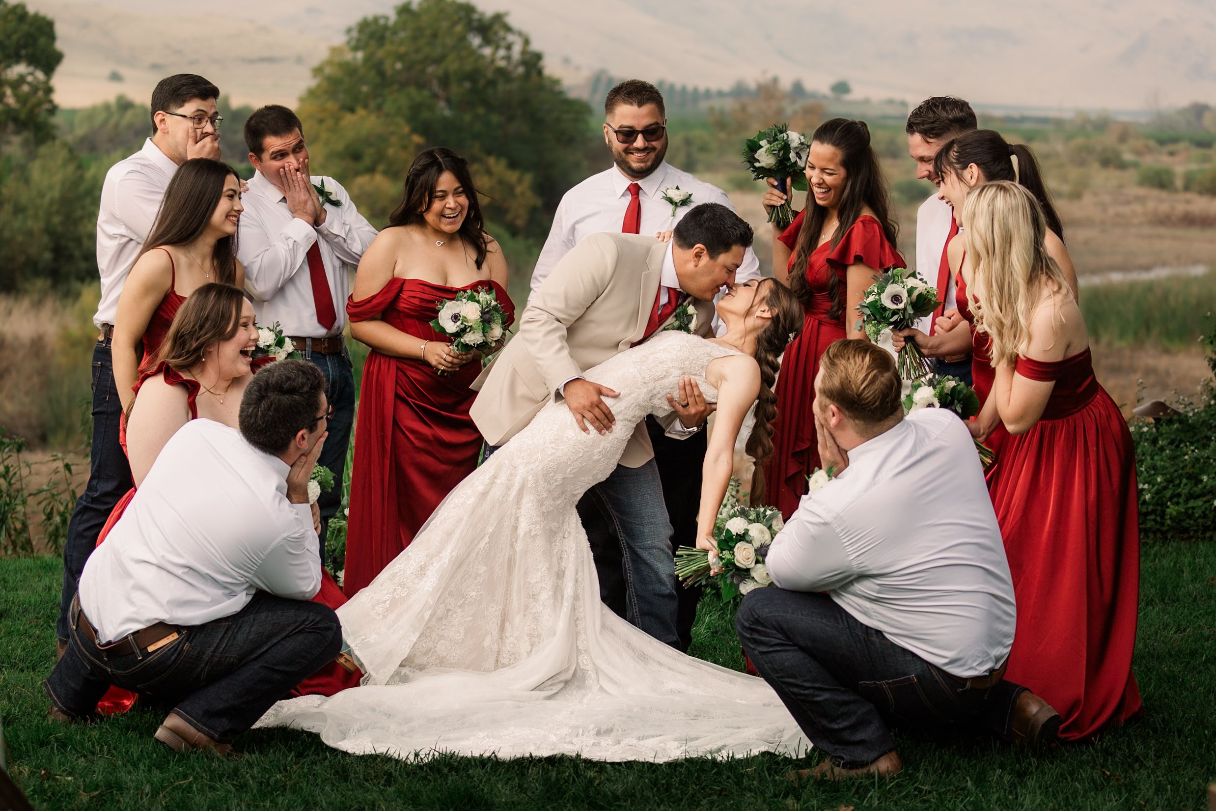 Wedding party in red attire relaxes on grassy hillside during sunset photo session.
