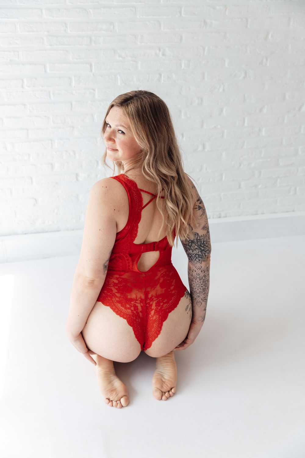 A woman in red lace lingerie poses against a white wall, showing a floral arm tattoo and wavy blonde hair.