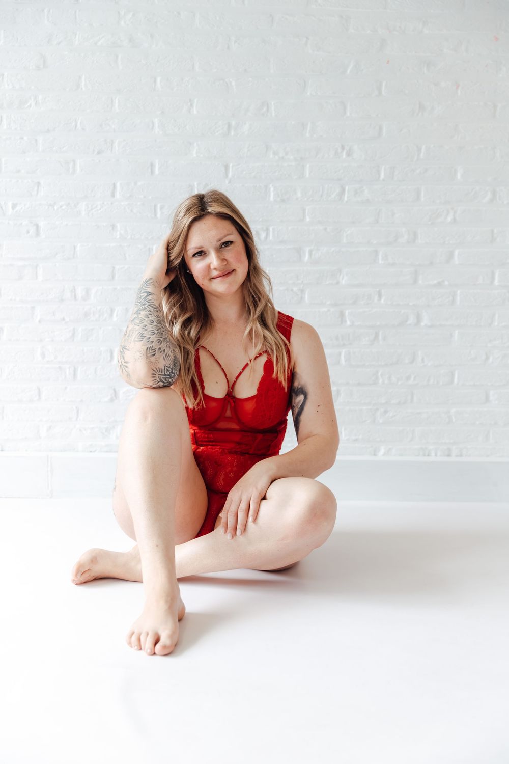 A woman in a red velvet bodysuit poses seated on a white floor against a white brick wall.