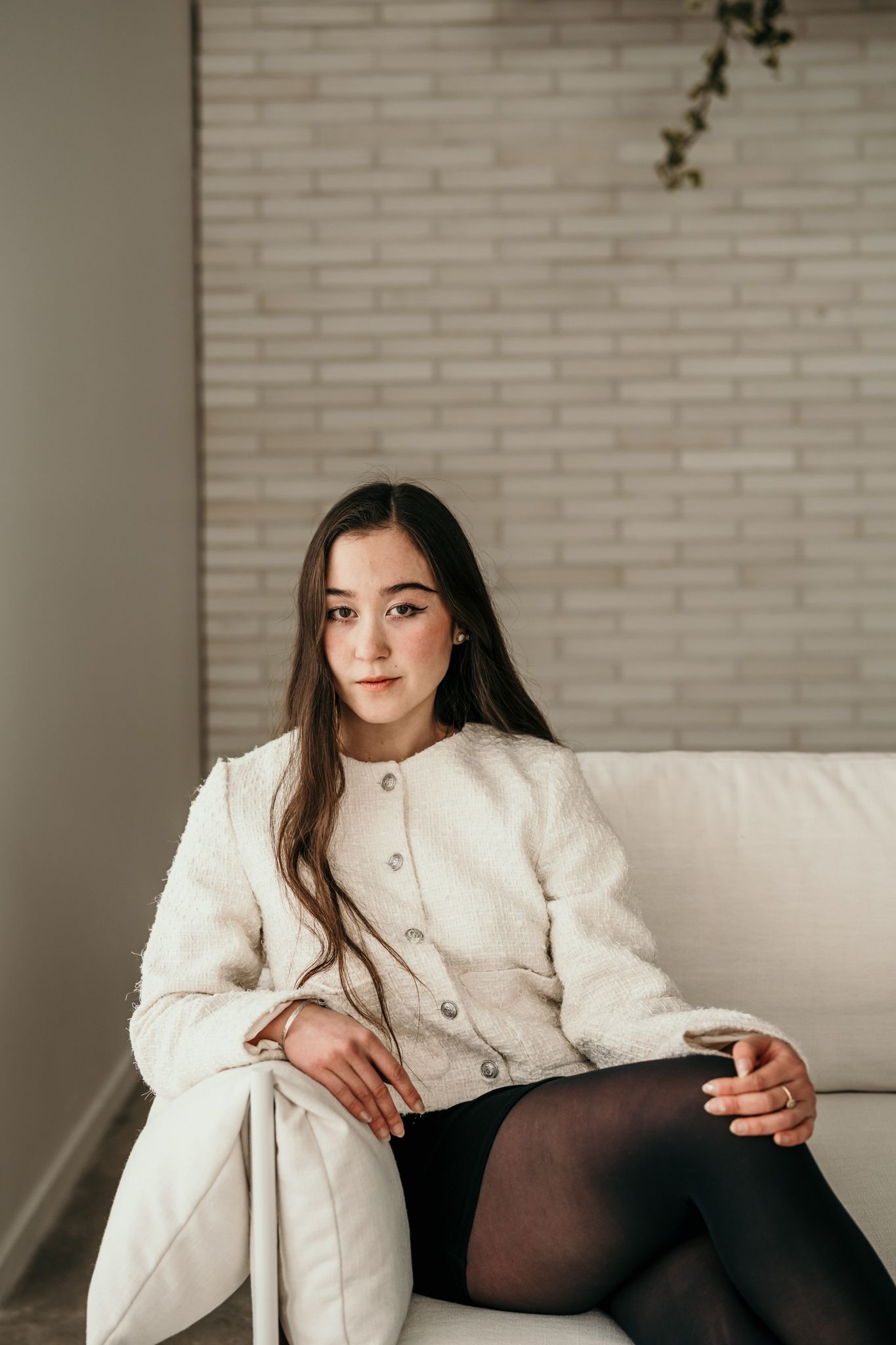 Person in white textured coat and black tights sits elegantly on cream colored couch against brick wall.