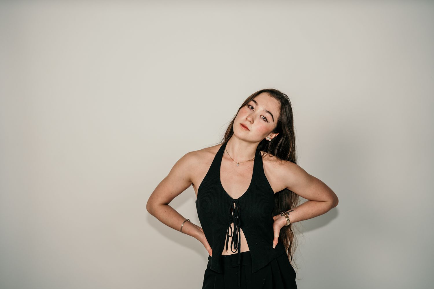 Woman in black sleeveless outfit poses against a light colored wall with a contemplative expression.