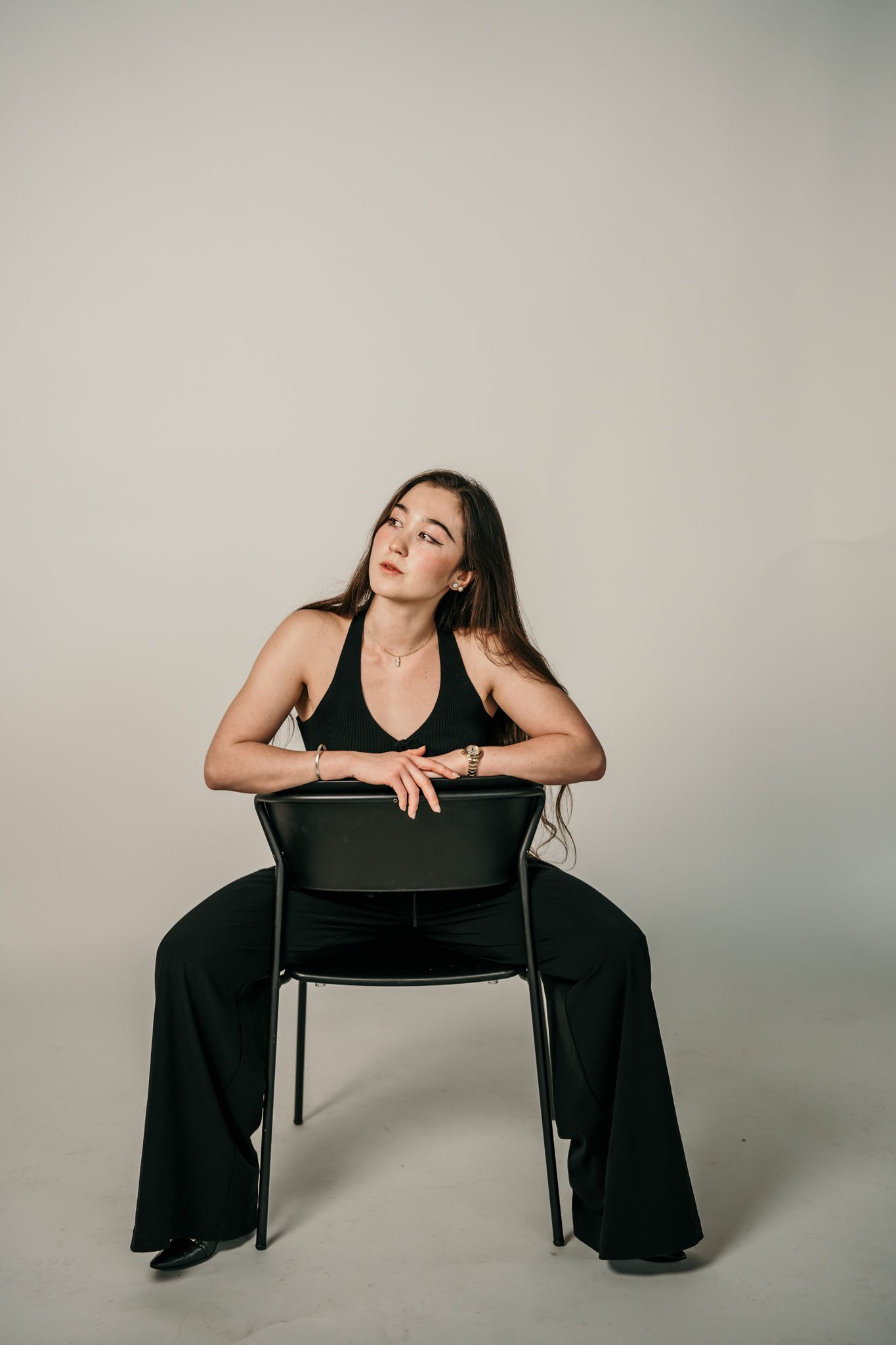 A person in black attire strikes artistic poses against a gray backdrop while interacting with a dark wooden chair.