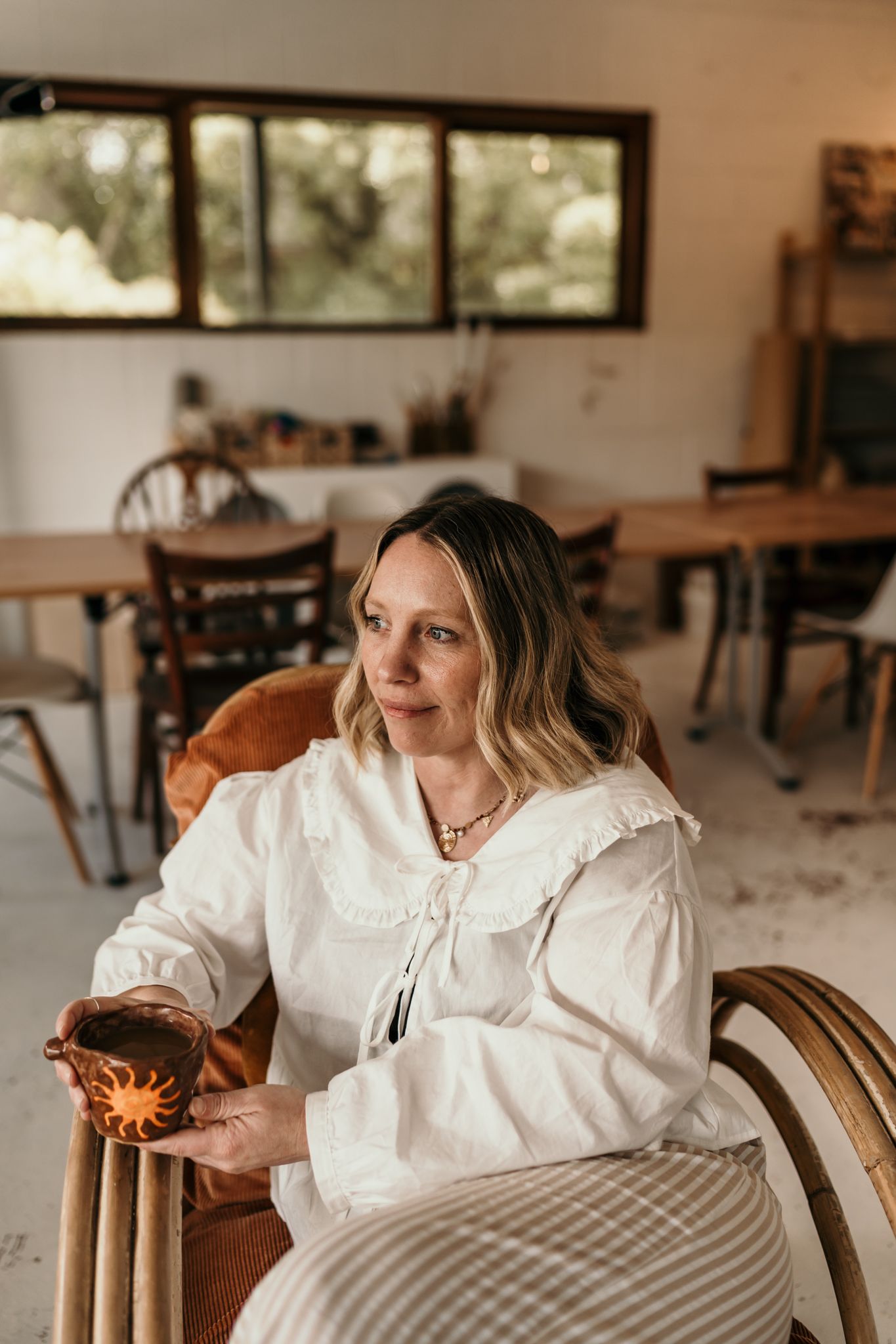 A person in a white dress sits in a cozy cottage interior holding a warm beverage in Christchurch pottery studio.