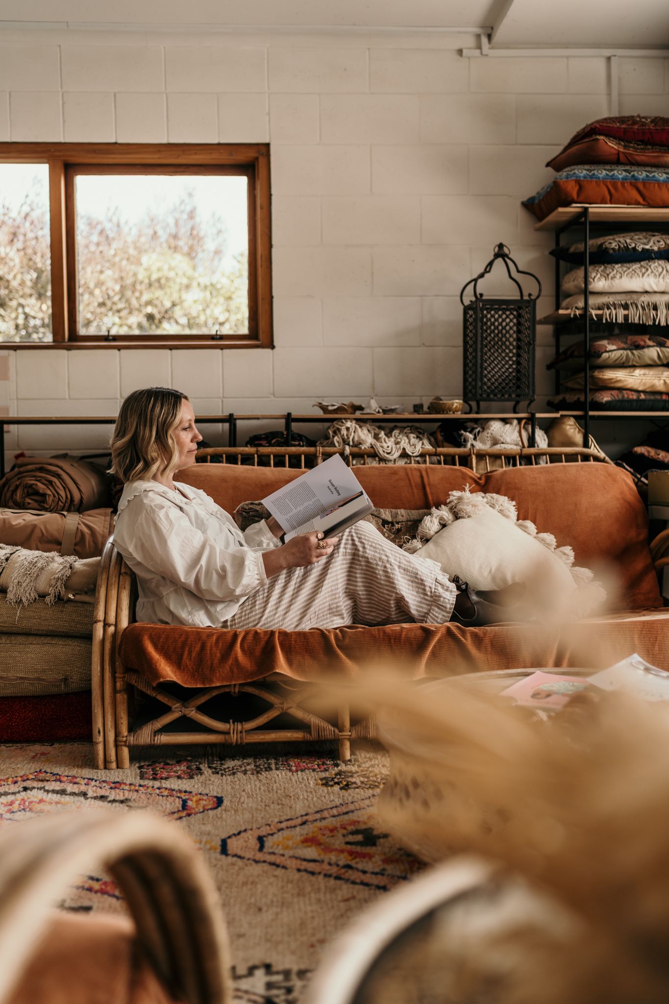 A person in white lounge on a rattan sofa while reading a book in a sunlit room in Christchurch pottery studio.