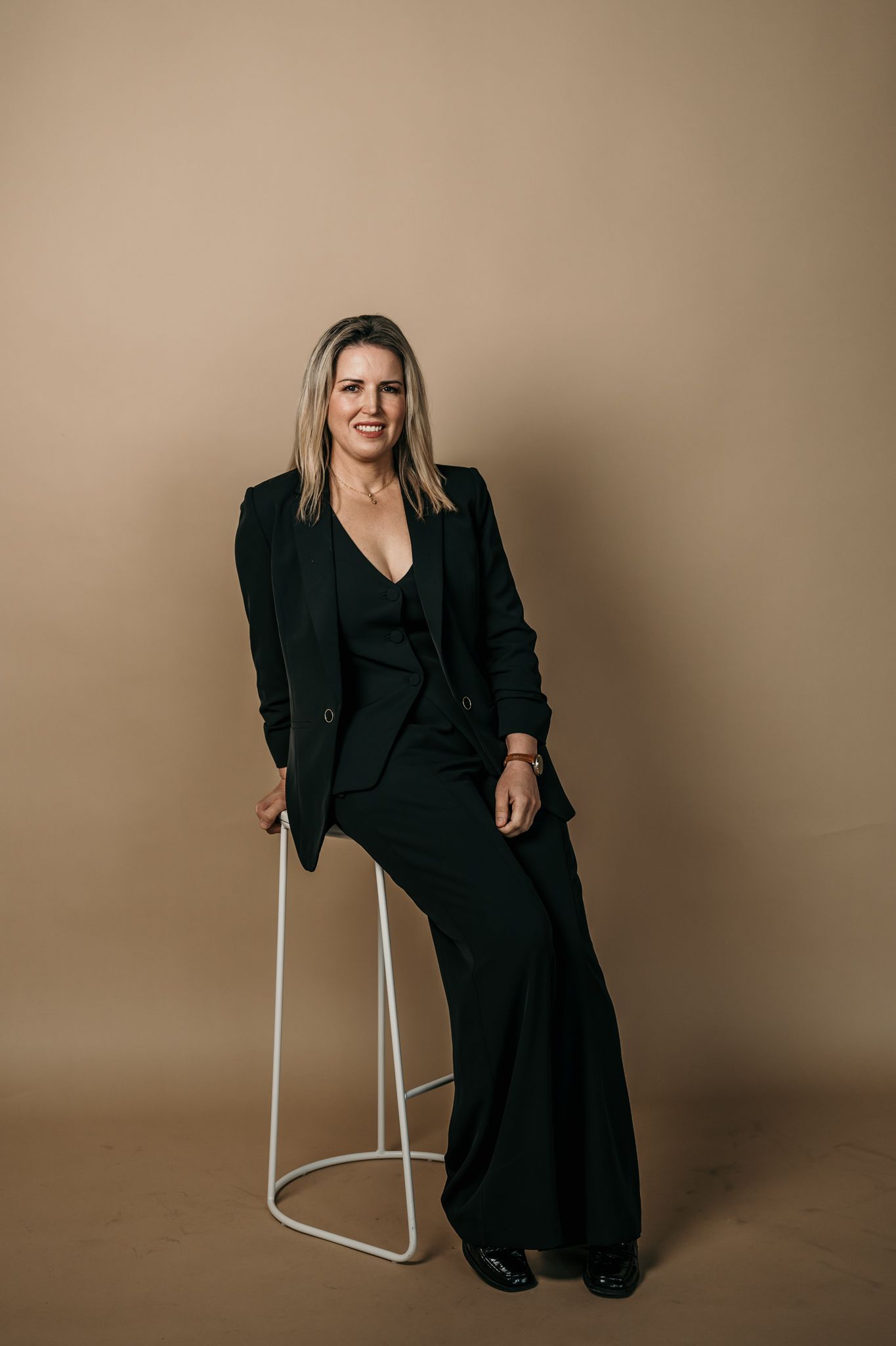 Professional figure in a black blazer and pants ensemble sits casually on white stools against a neutral backdrop.
