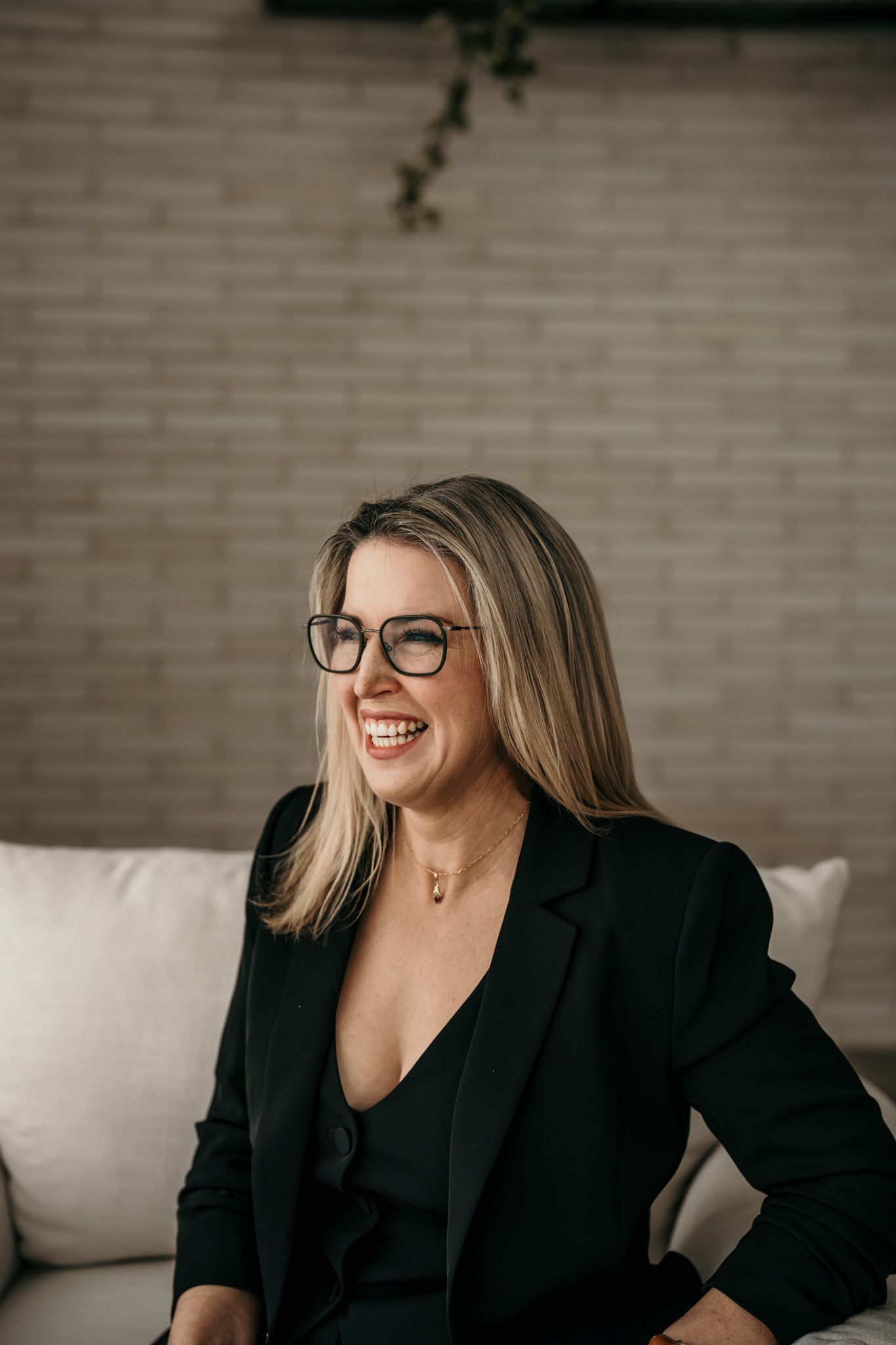 Professional wearing glasses and a black blazer sits relaxed against white cushions while laughing joyfully.