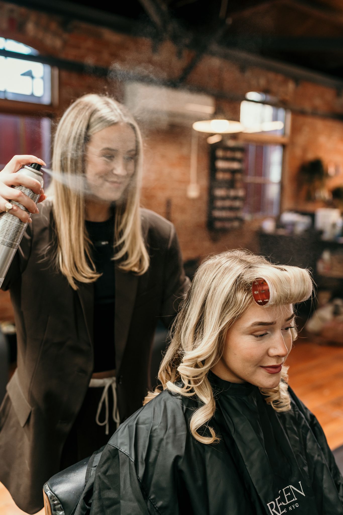 Hairstylist applying hairspray while styling client's blonde curls in vintage-inspired look at rustic salon.