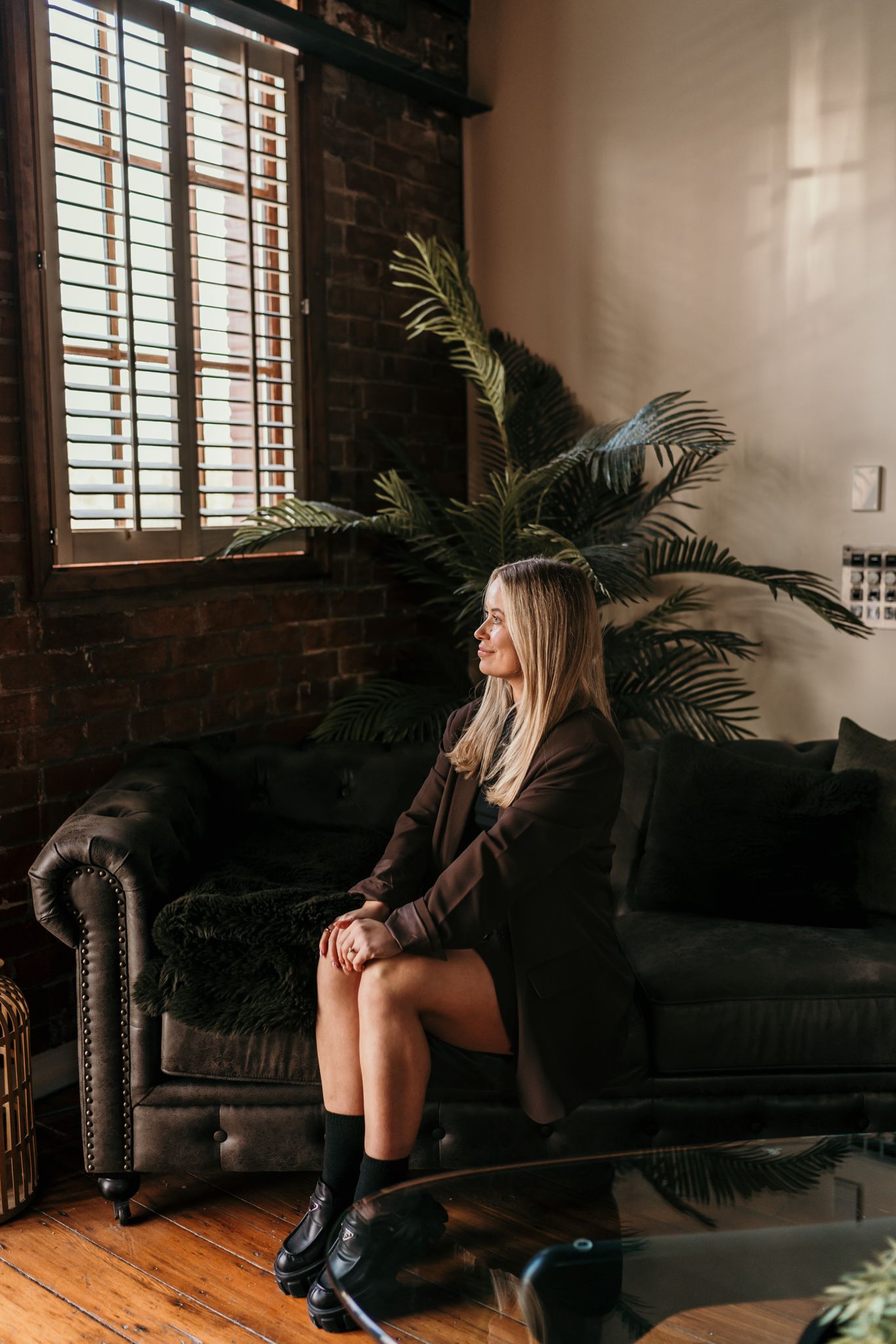 Three moody boudoir shots show someone posing in silhouette on a leather couch near a potted palm in a loft space.