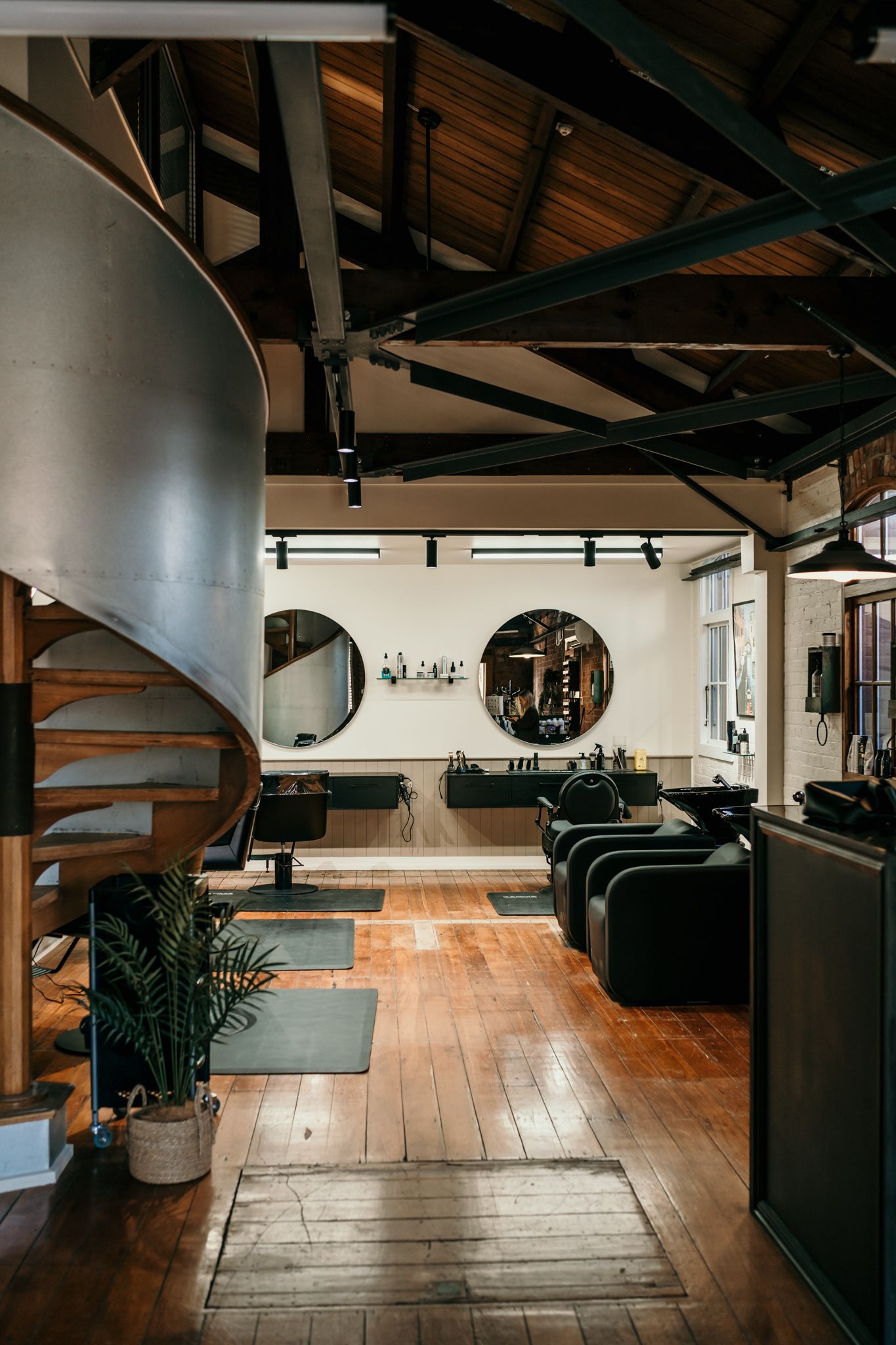 Modern salon interior with dark wood floors, spiral staircase, round mirrors and exposed beam ceiling in rustic industrial style.