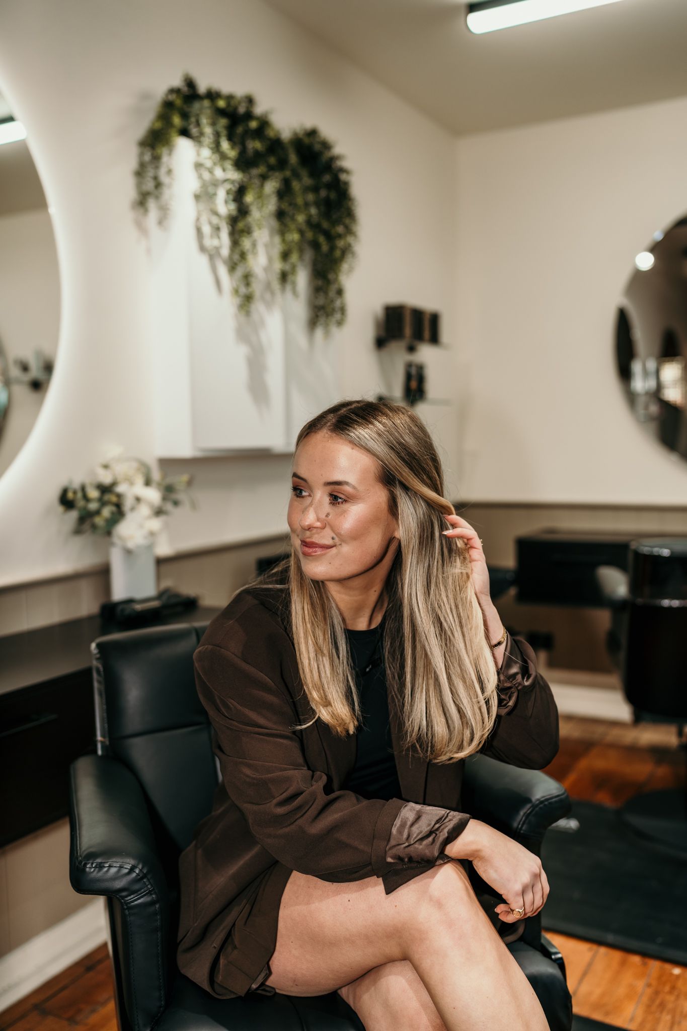 A person in a brown jacket sits casually on a black leather chair in a salon with hanging plants and hardwood floors.