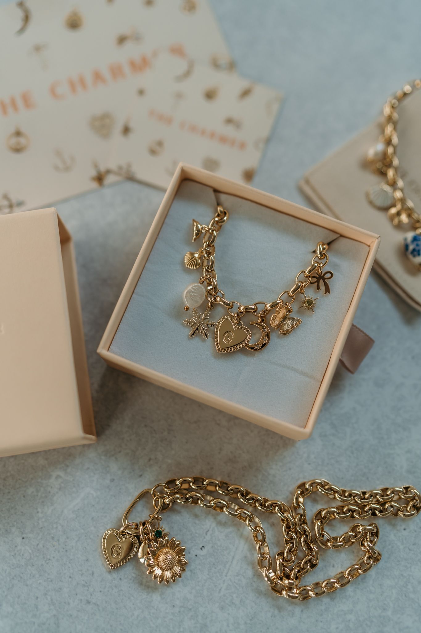Gold charm bracelet with nautical themed charms displayed in a pink box with additional jewelry pieces nearby.