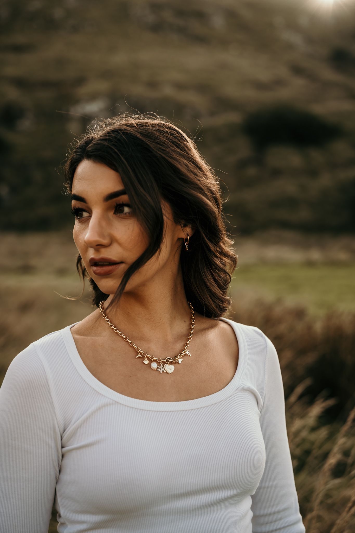 Portrait of a person with dark hair wearing a white long sleeve top and jewelry in a natural outdoor setting at sunset.