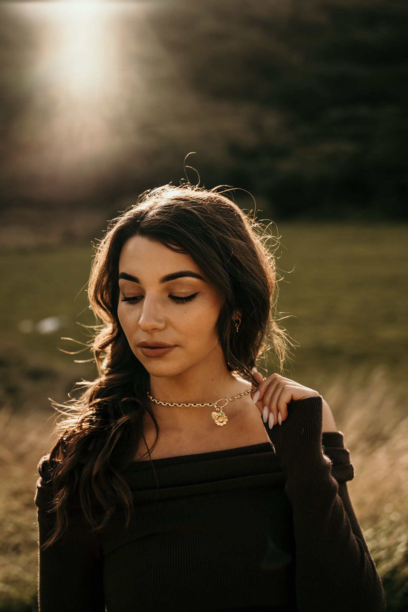 Portrait at golden hour with glowing sunlight and delicate necklace detail in a dark outfit with natural outdoor setting.