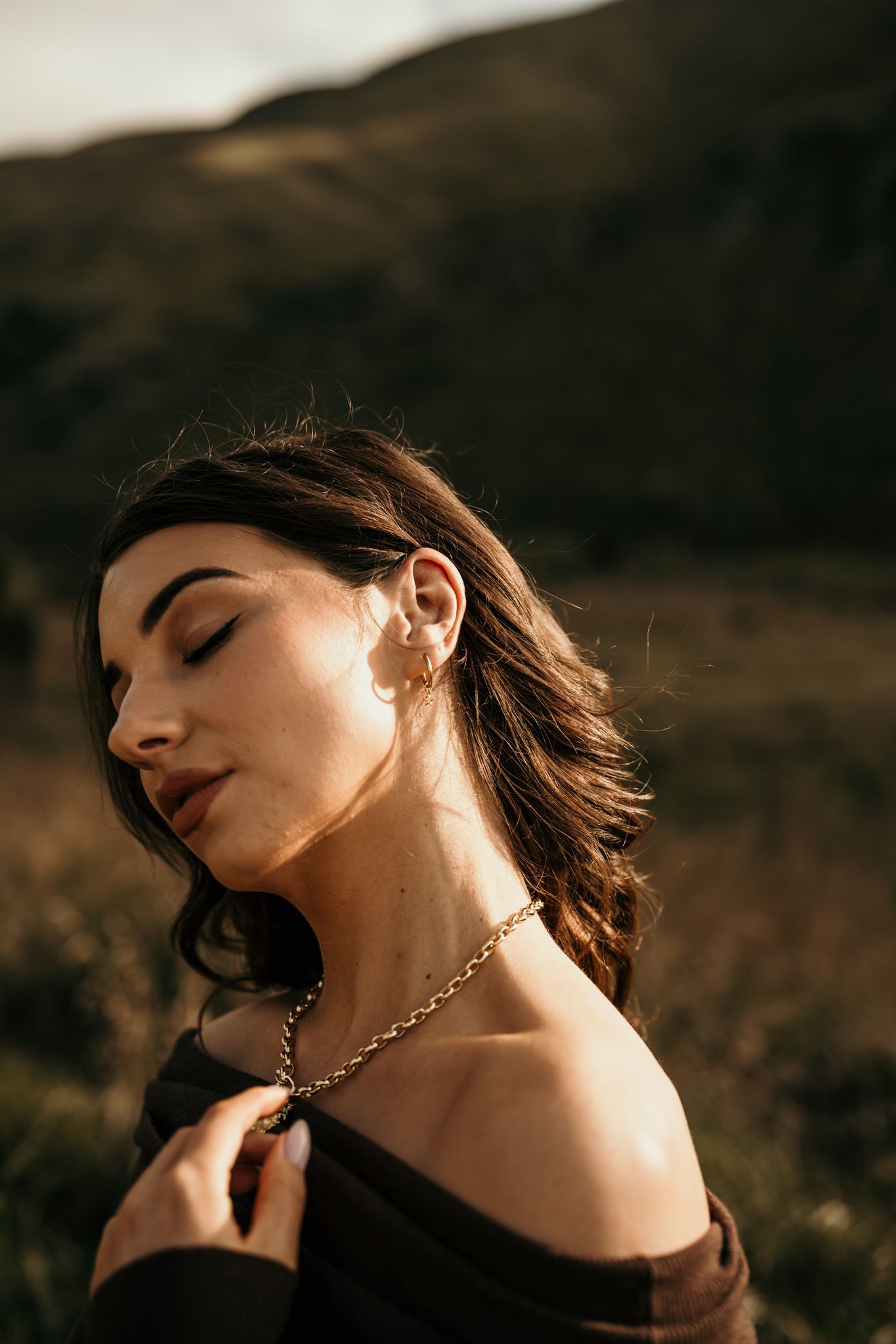 Golden sunlight illuminates figure in black off-shoulder top with gold necklace against mountainous backdrop.
