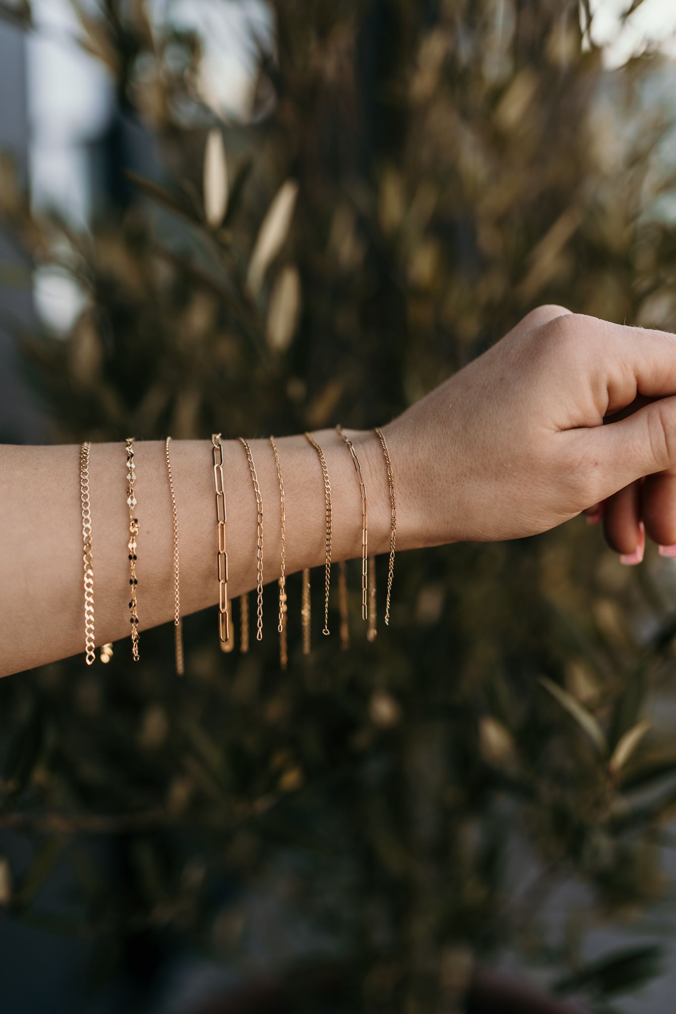 Multiple delicate gold chain bracelets displayed on an extended arm against a blurred natural background.