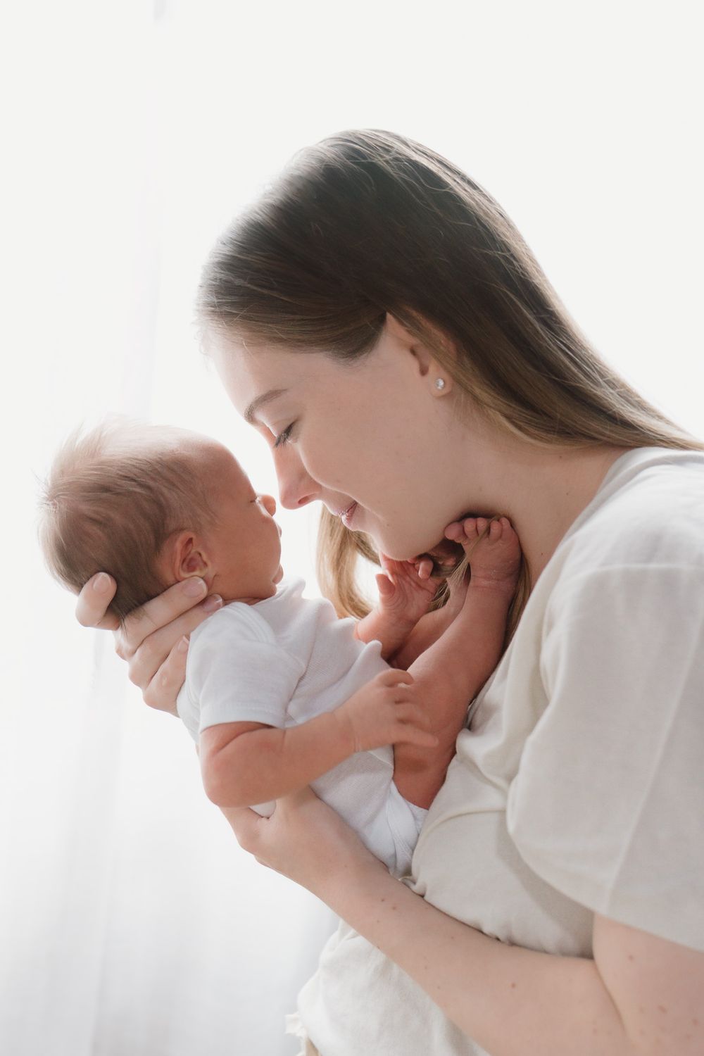 A woman is standing in front of a white backdrop and touching her nose to her sleeping babies nose