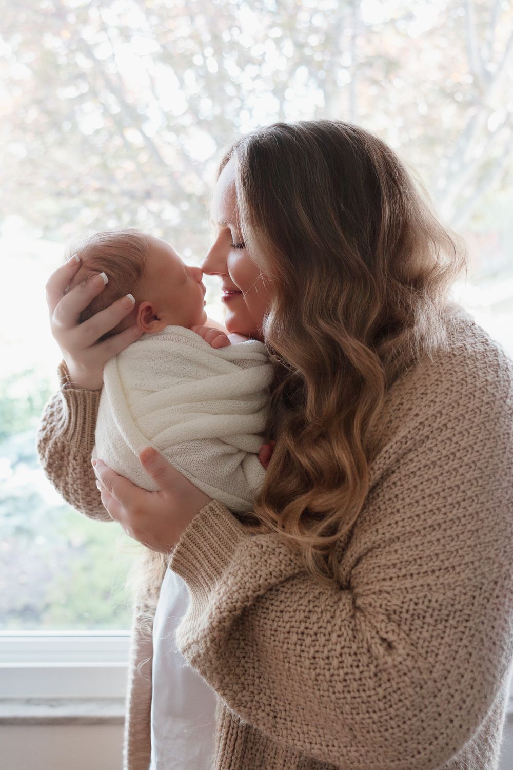 A woman is standing in front of a window and touching her nose to her sleeping babies nose