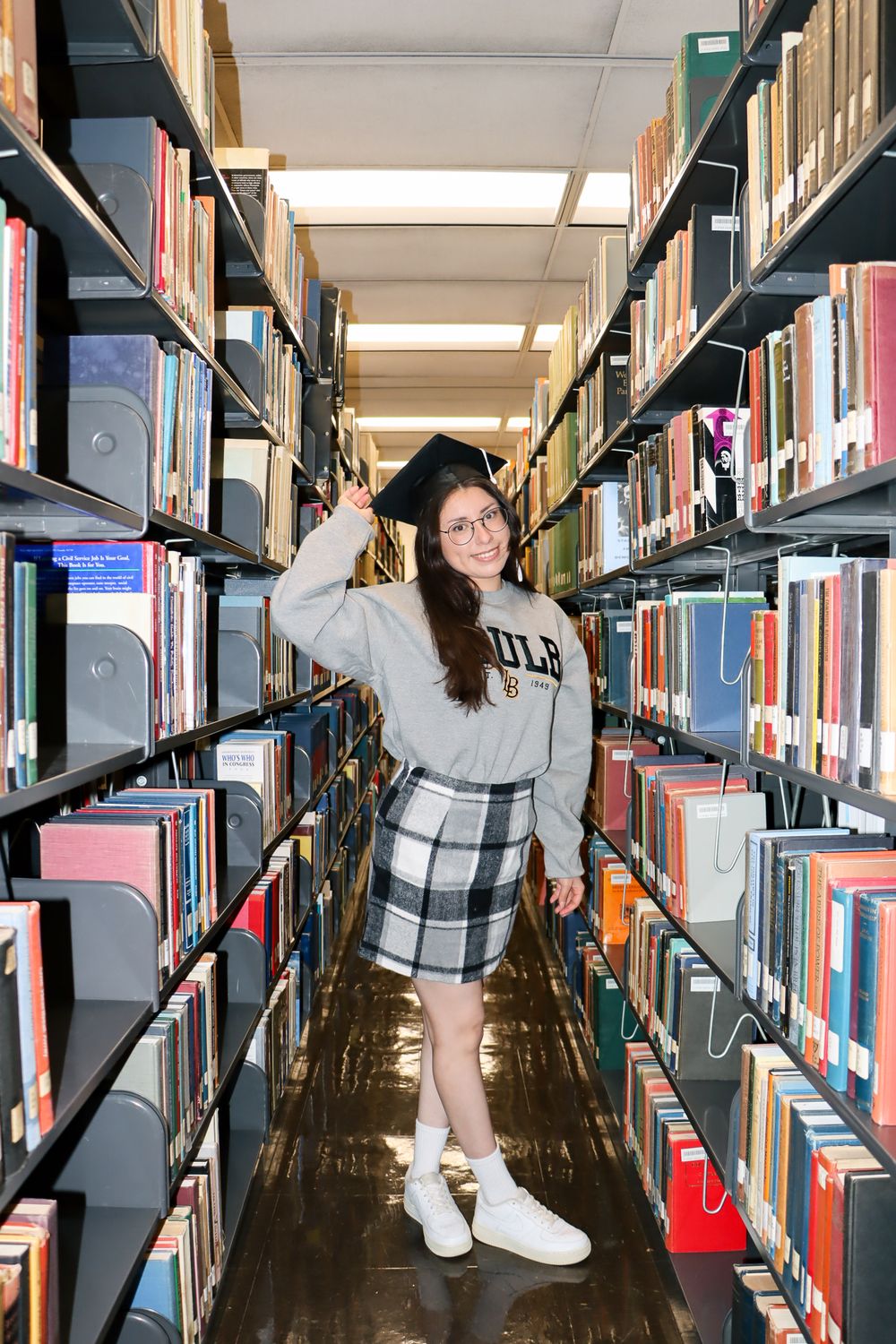 Graduate poses playfully between library bookshelves wearing casual outfit and graduation cap.