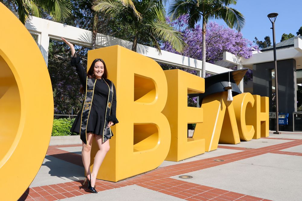A graduate poses next to large yellow BEACH letters in an outdoor setting with palm trees.