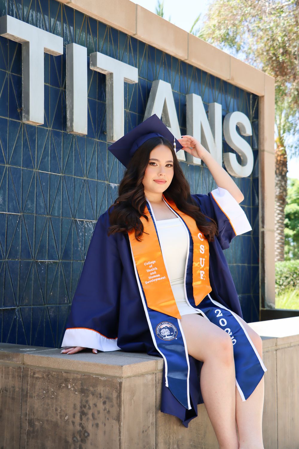 Graduate in academic regalia poses against Titans building wall.