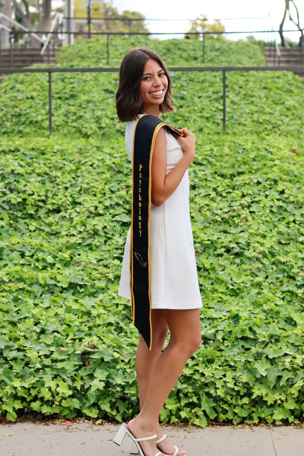 Graduate in white dress poses with graduation stole against green ivy covered wall.
