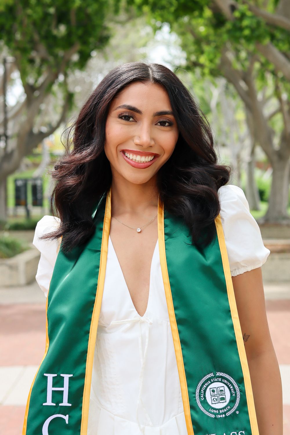 Graduate wearing white dress and green graduation stole poses in front of tree-lined walkway.