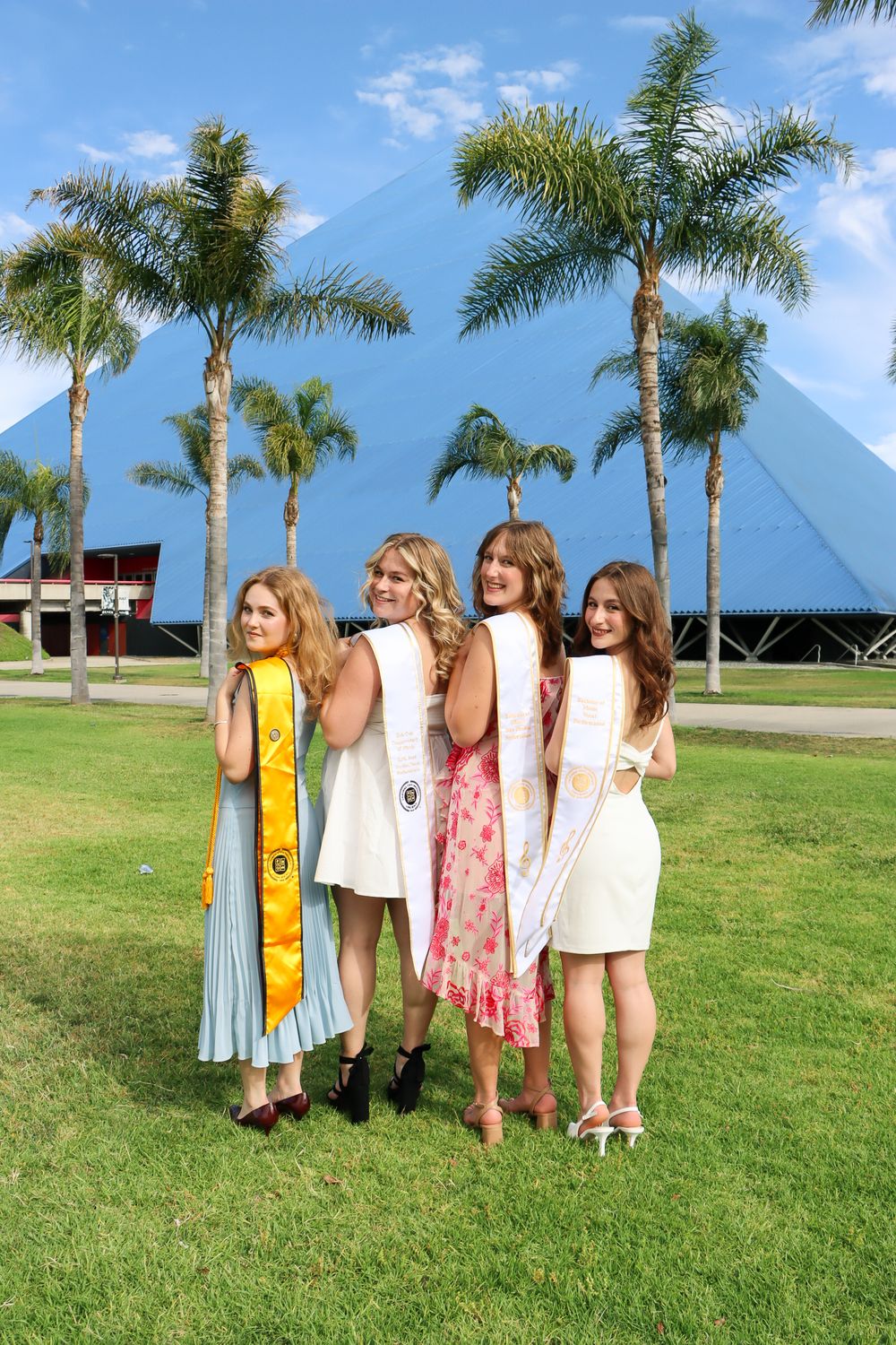Four graduates in light dresses pose together in front of a blue pyramid structure with palm trees.
