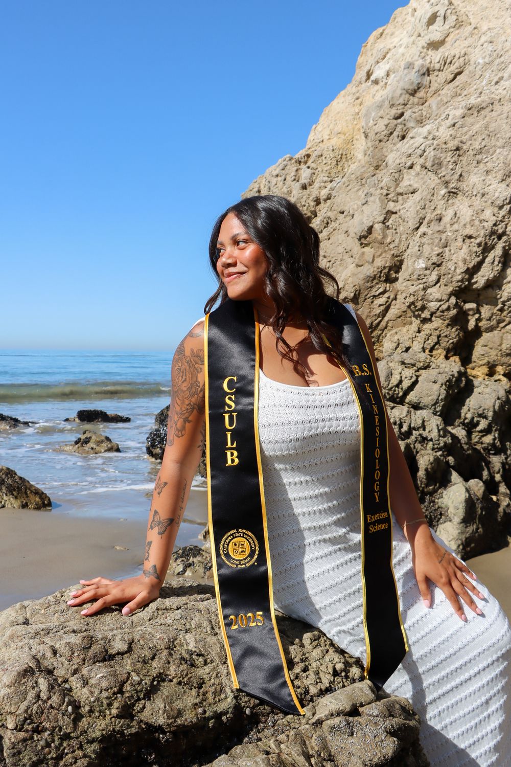 Graduate in white dress and academic stole poses on rocky beach with ocean view.