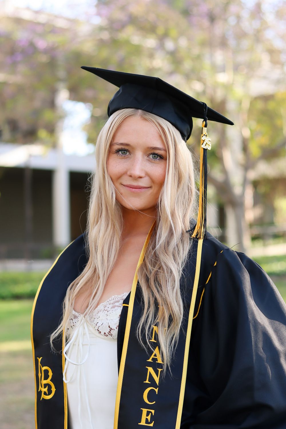 Graduate wearing black graduation gown with gold honor cords against campus backdrop.
