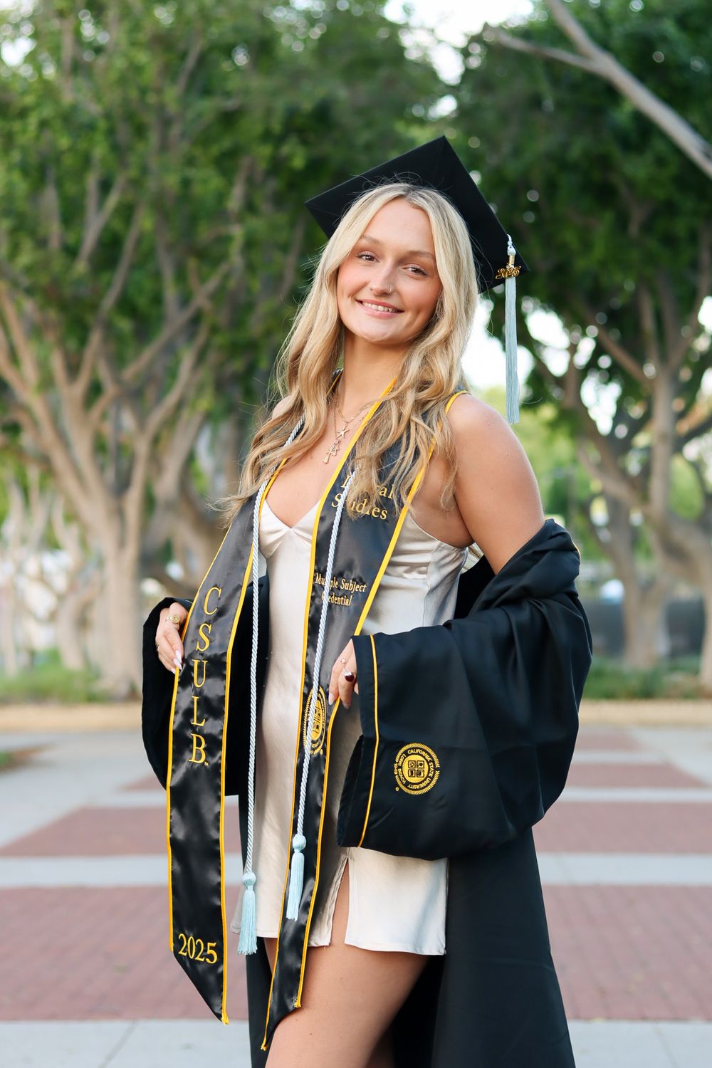 Graduate in white dress and black graduation gown poses outdoors on brick pathway.