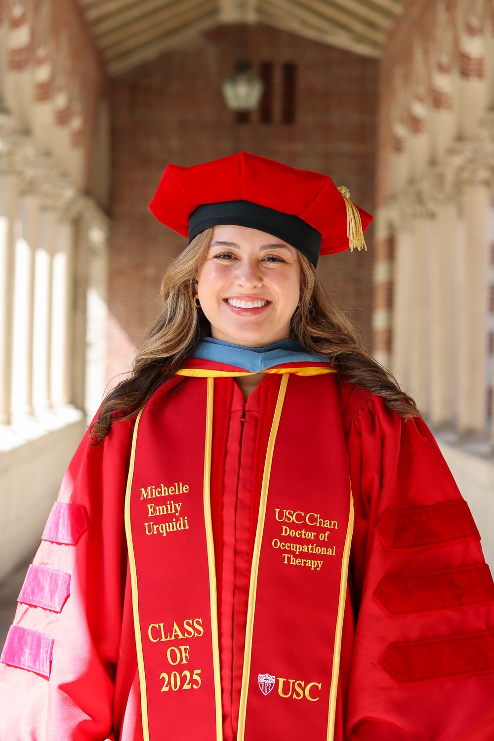 Graduate in red doctoral regalia poses in university colonnade.